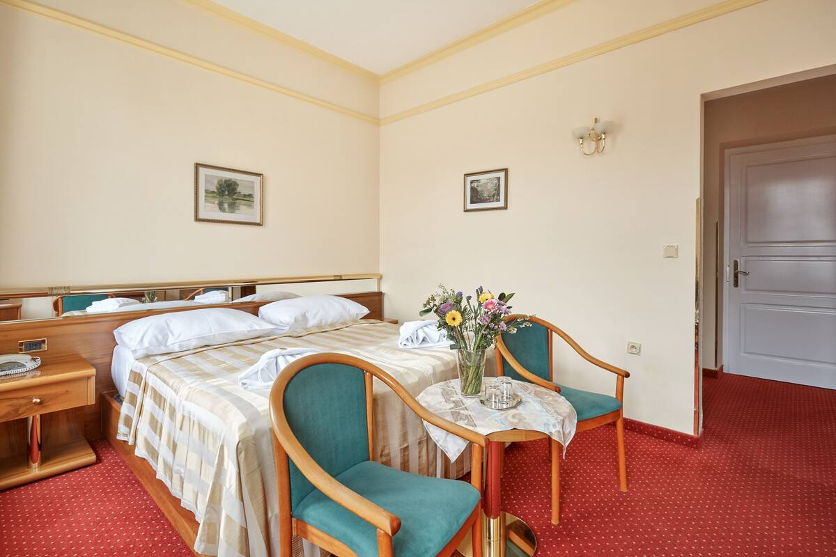 Una habitación de hotel con una cama, dos sillas, una mesita con un florero y una puerta.