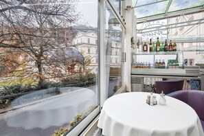 Ein Restaurant mit Blick auf eine von Bäumen gesäumte Straße und Gebäude. Ein Tisch ist mit Salz- und Pfefferstreuern sowie einer Wasserflasche gedeckt.