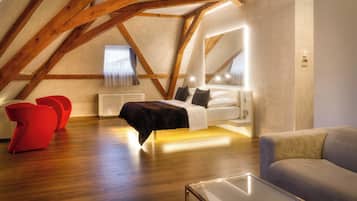 A modern loft bedroom with a sloped ceiling, wooden beams, a bed, red chairs, and a gray sofa.