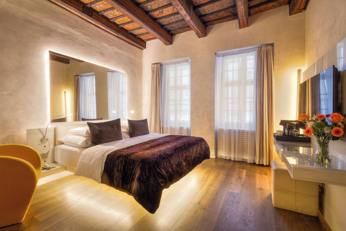 Une chambre à coucher moderne avec un grand lit, une table de chevet, un miroir et un bureau agrémenté d’un vase de fleurs.