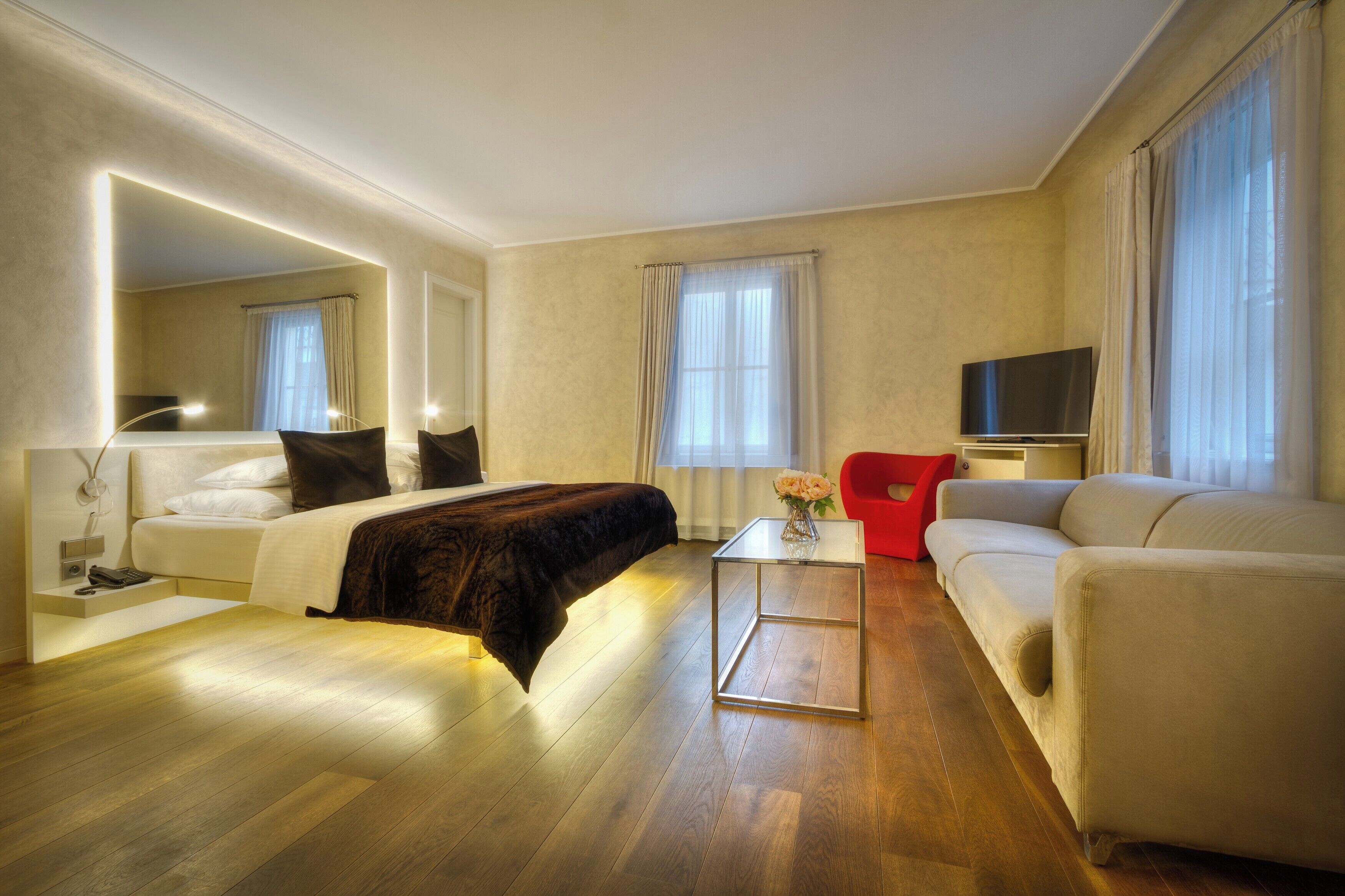 A modern hotel room with a bed, a sofa, a TV, and a small table with a vase of flowers.