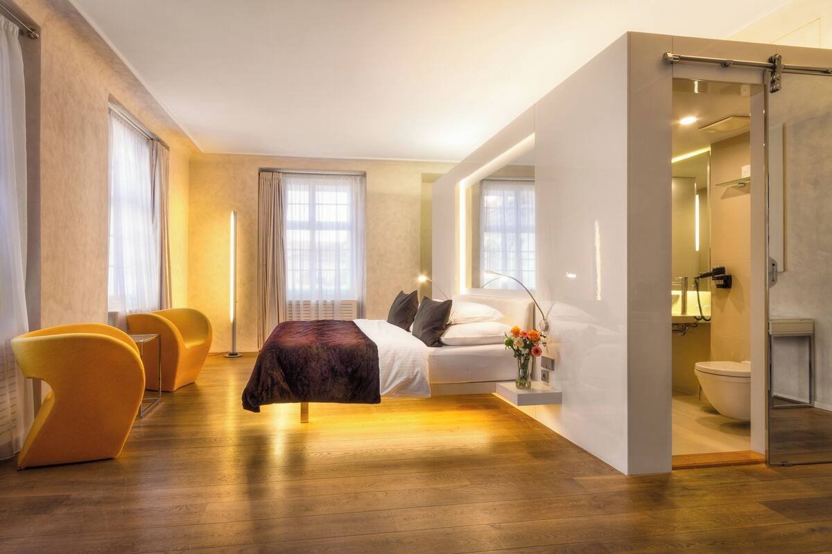 Une chambre d’hôtel moderne, équipée d’un lit, de deux fauteuils, d’une table de chevet, d’une lampe, d’un vase avec des fleurs et d’une salle de bain visible à travers une porte ouverte.