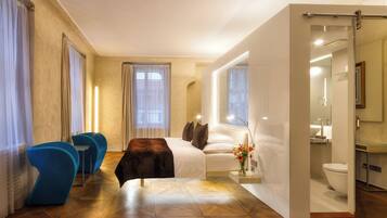 A modern hotel room with a large bed, a sitting area with blue chairs, a bathroom with a toilet and sink, and a view of the city.