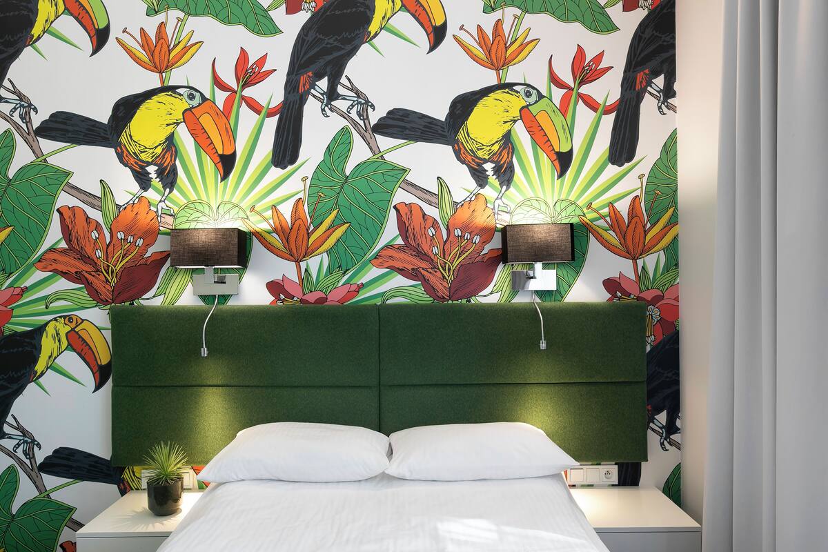 Et hotelværelse med en grøn polstret sengegavl, hvidt sengetøj og et vægmaleri med tukaner og tropiske blomster.