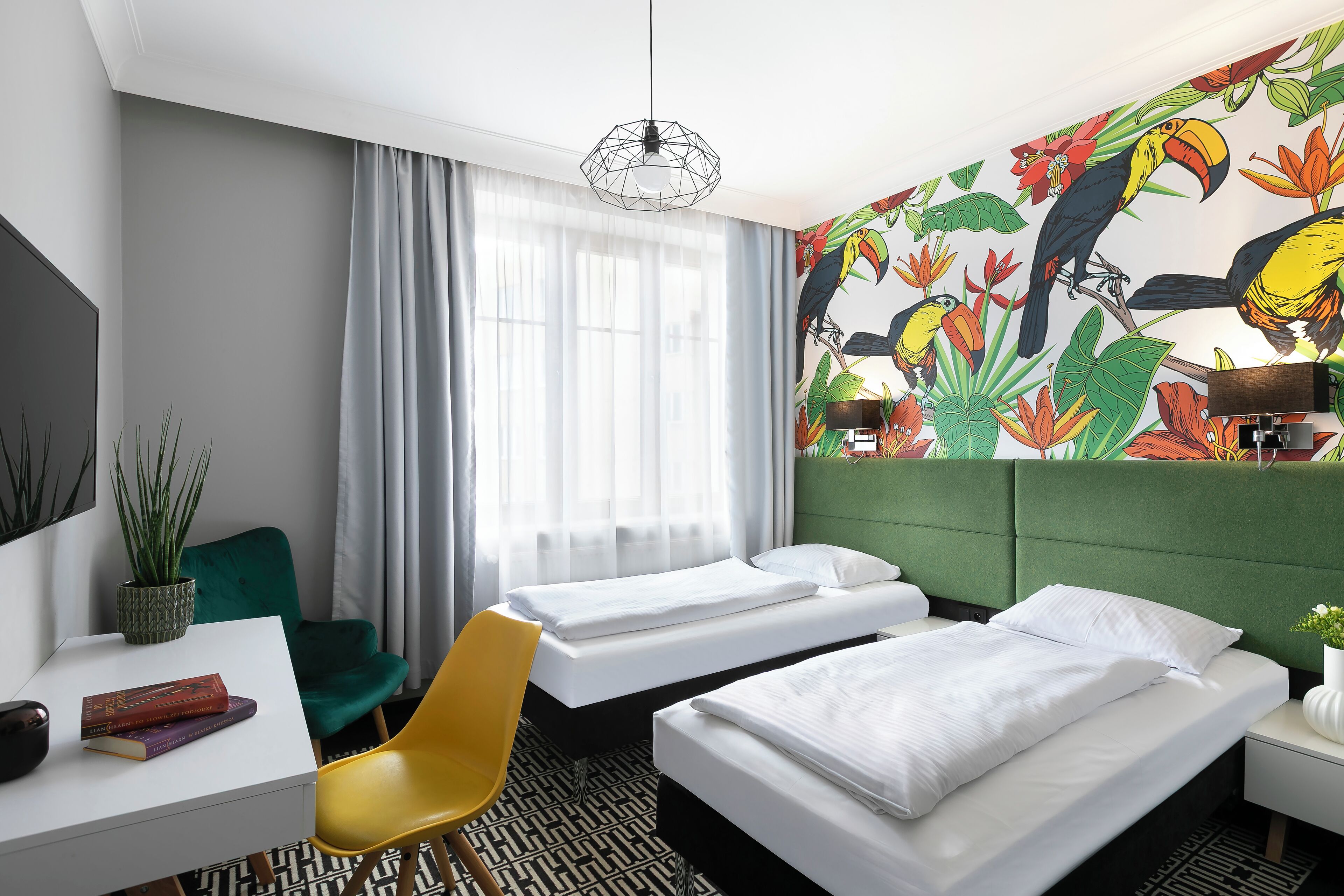 A hotel room with two beds, a desk, a chair, a potted plant, and a mural of toucans.