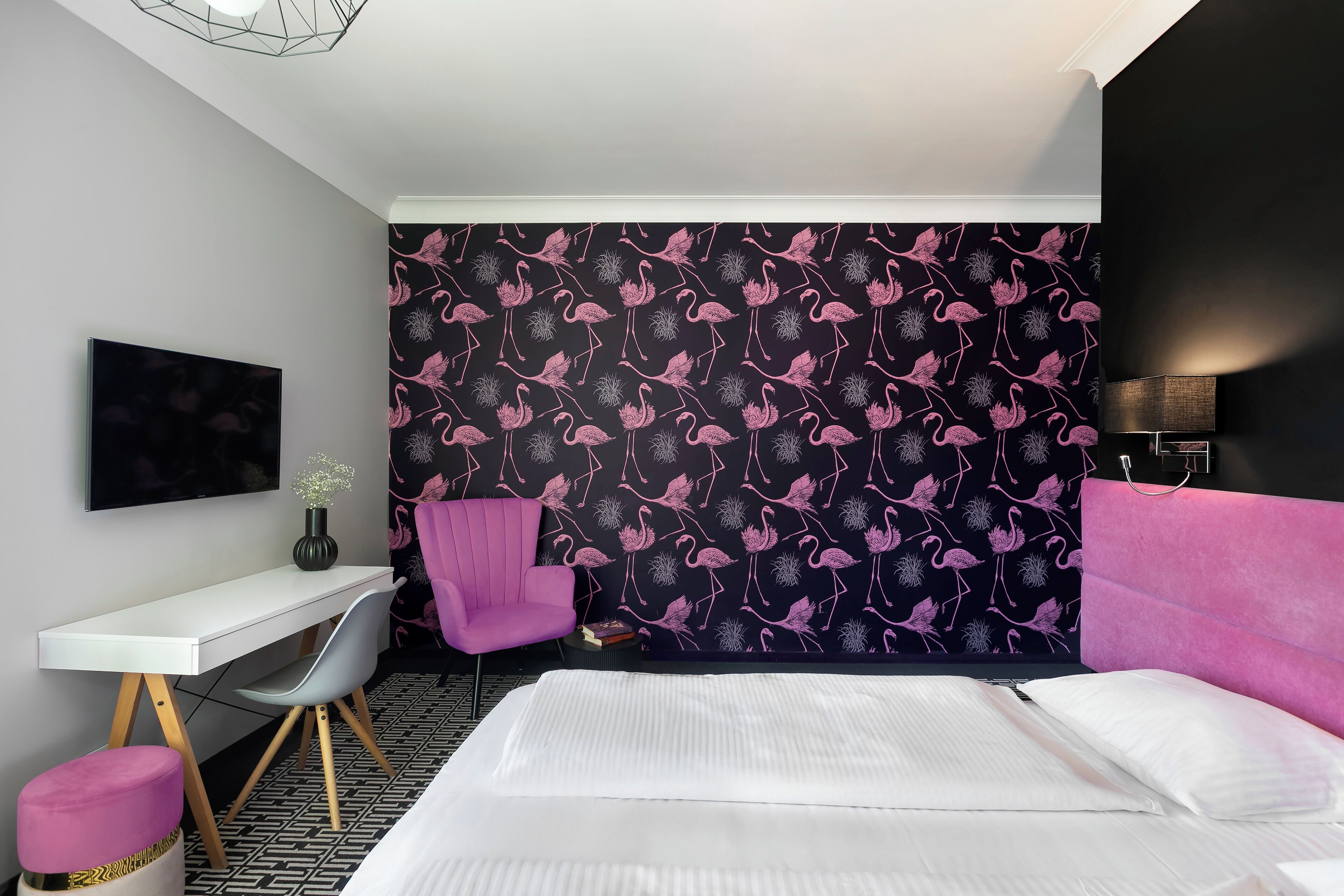 A modern hotel room with a large bed, a desk, a chair, and a wall with a black and pink flamingo pattern.