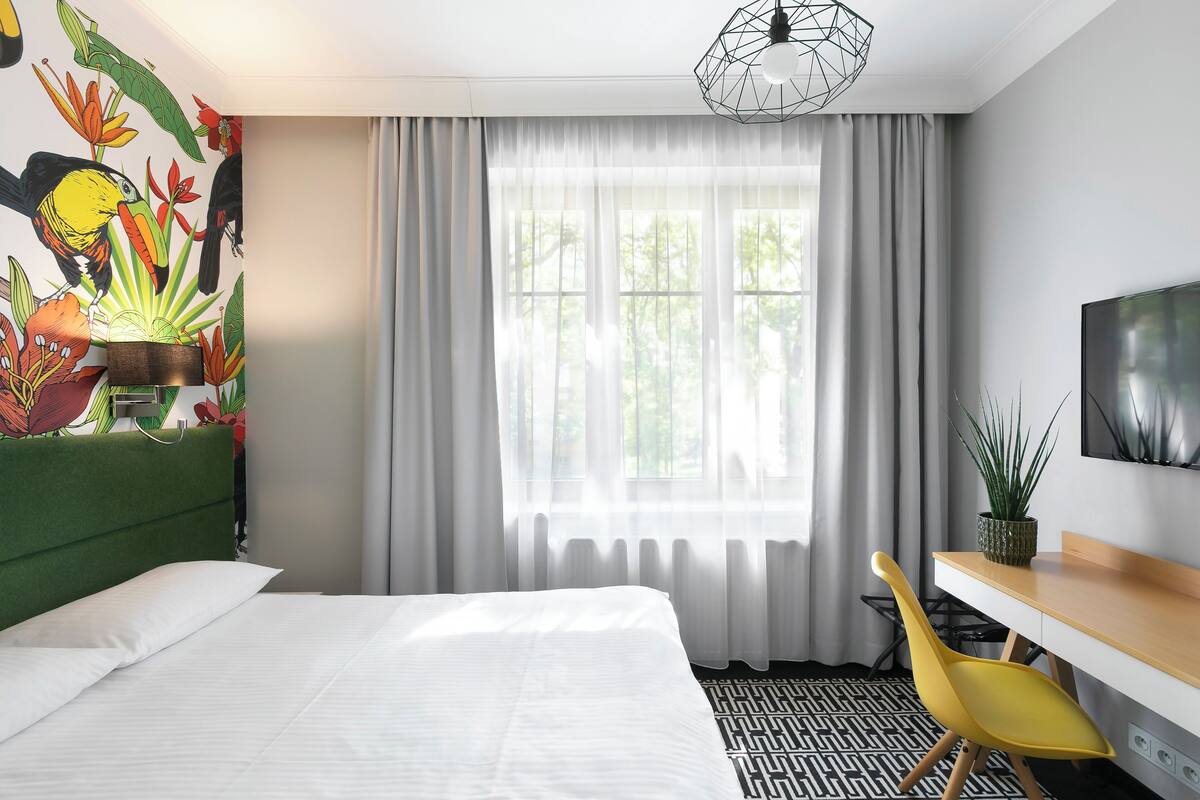 Et hotelværelse med seng, skrivebord med stol, fjernsyn og et vindue med gardiner.