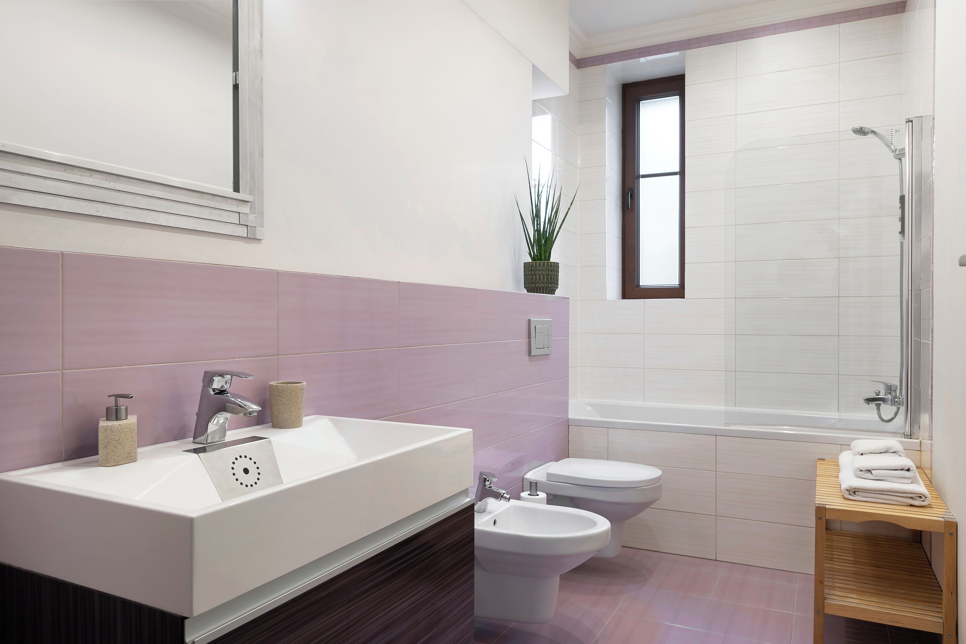 A modern bathroom with a large sink, two toilets, a shower, and a small wooden stool with towels.