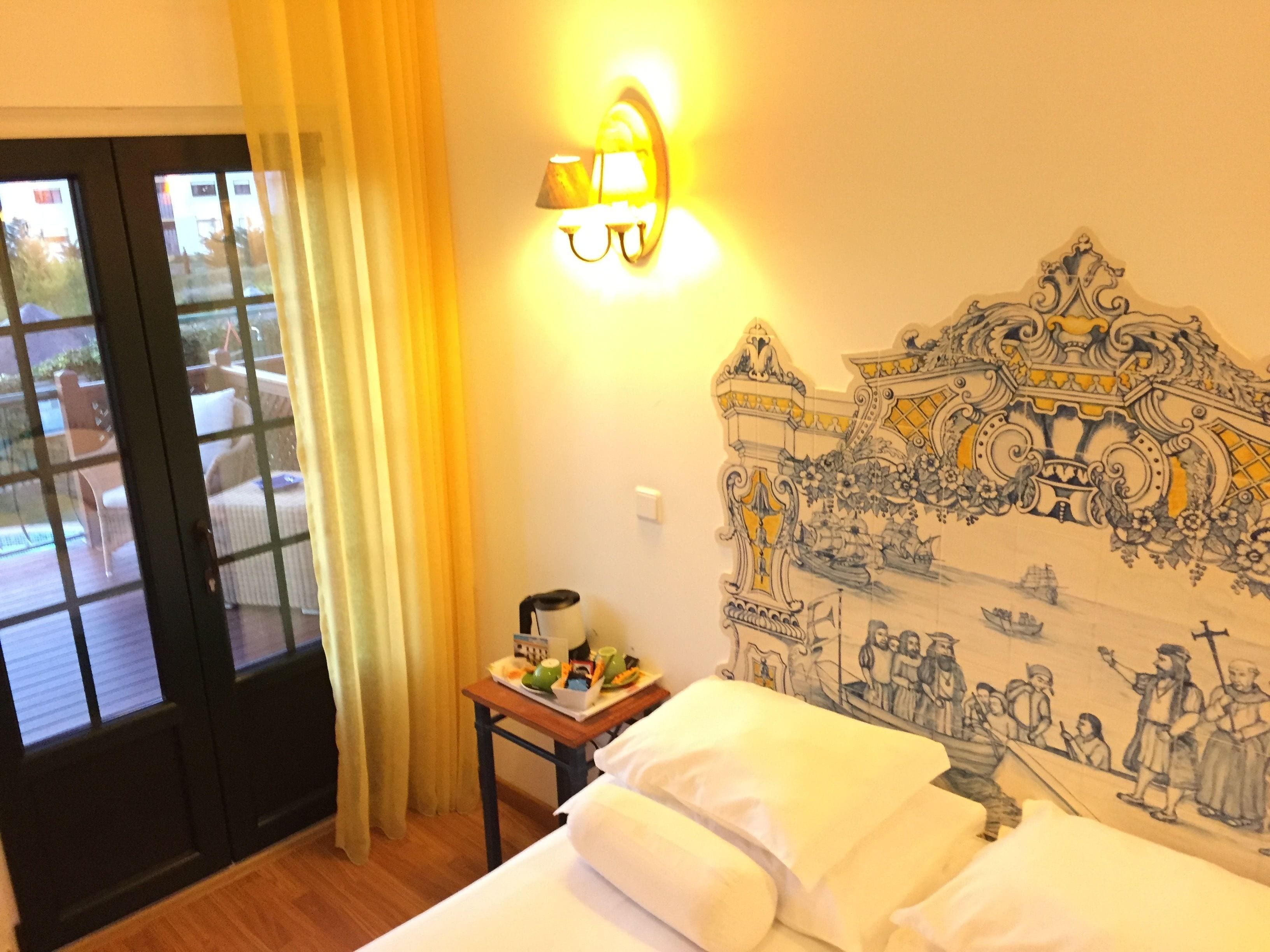 A hotel room with a bed, a nightstand with a lamp, a wall mural, and a view through a door.