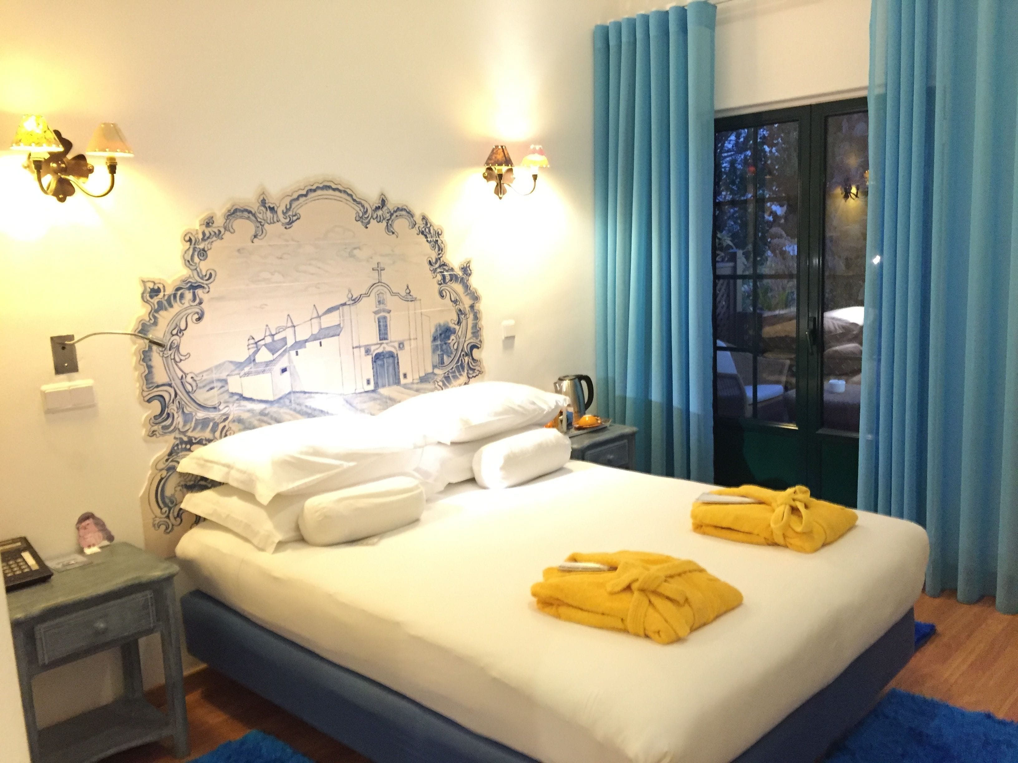 Habitación de hotel con cama, mesita de noche, lámpara, un cuadro y vistas al exterior.