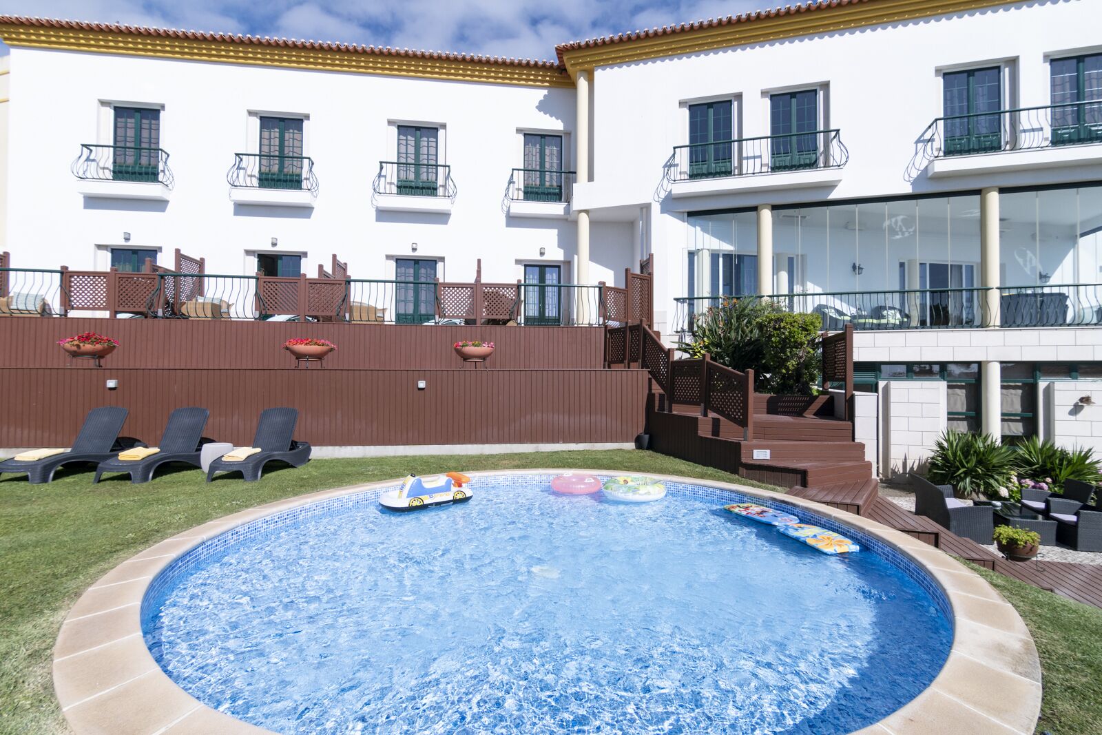 A swimming pool with lounge chairs, a wooden deck, and a white building with balconies.