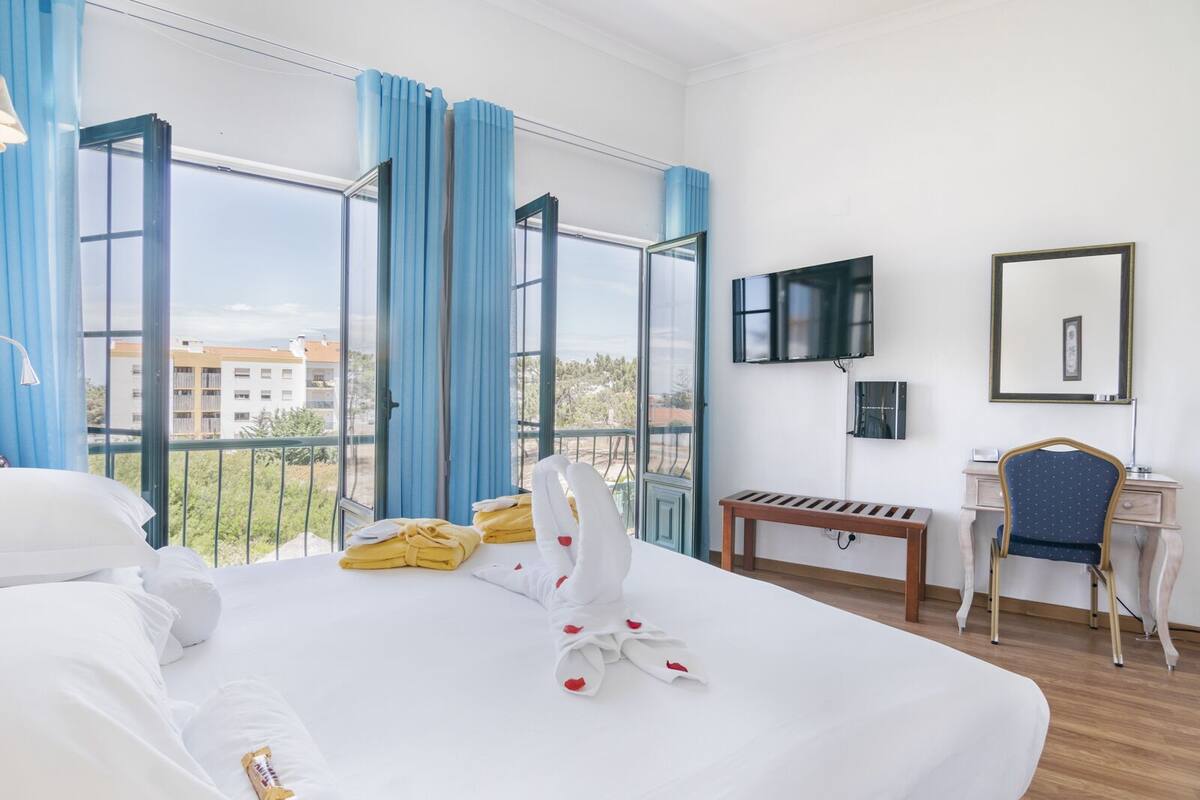 A hotel room with a large bed, a desk, a chair, a TV, and a balcony with blue curtains.