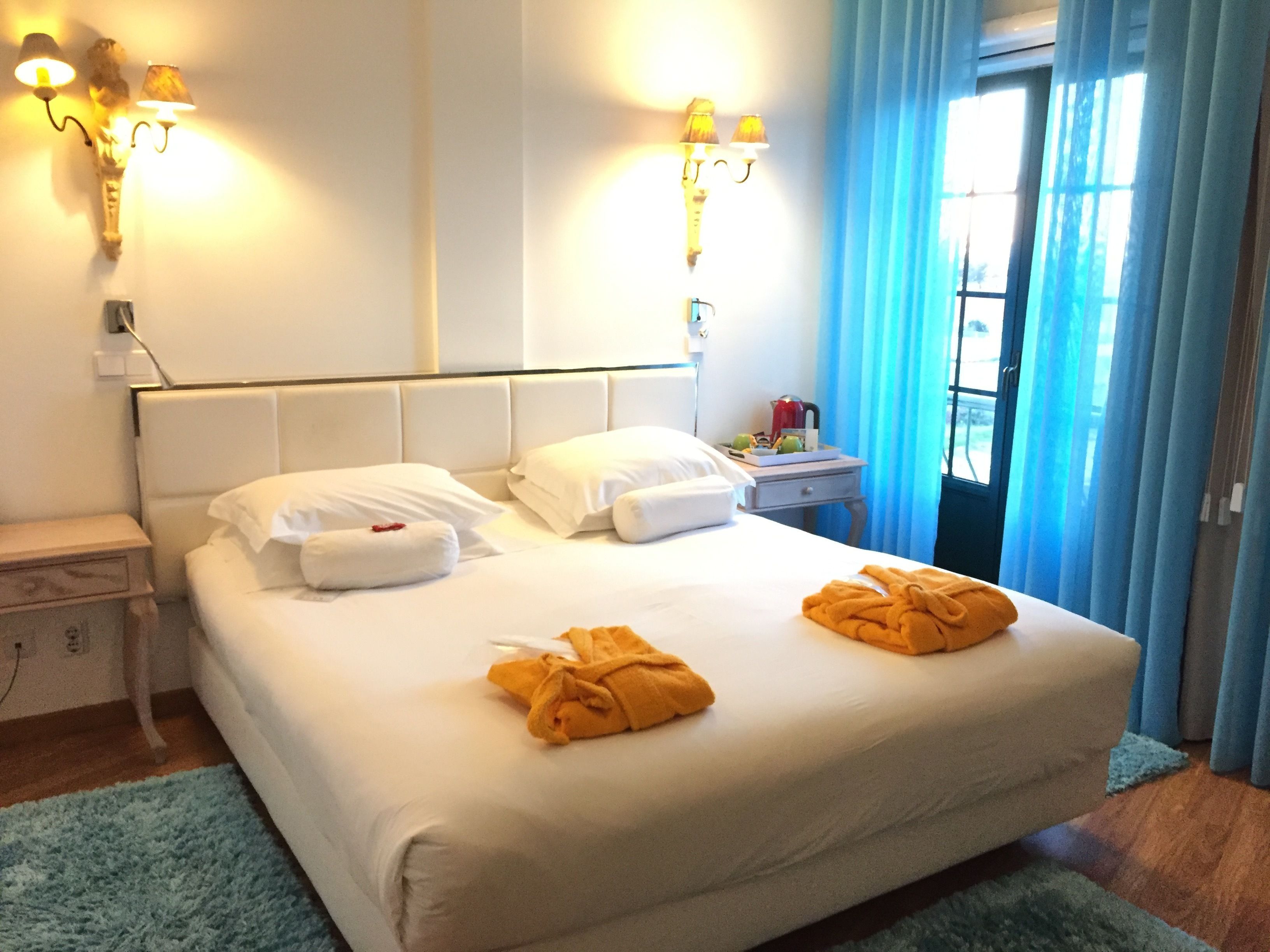 A hotel room with a bed, two bedside tables, a nightstand, a window with curtains, and wall-mounted lighting.