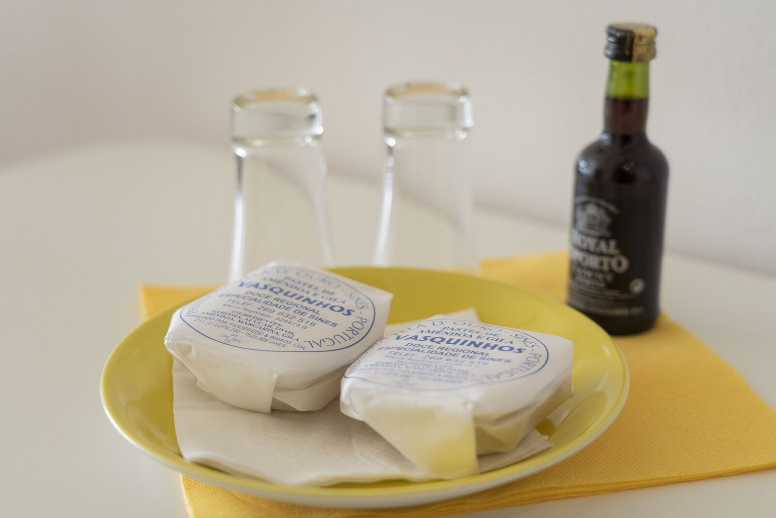 Un plato con dos productos envueltos, etiquetados como 'VASQUINHOS', y una botella de vino 'Royal Porto' sobre una servilleta amarilla.