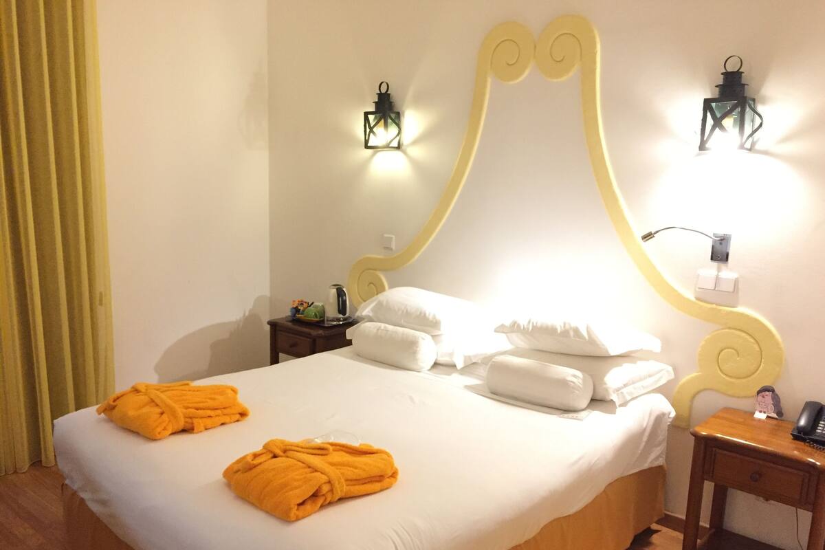 A hotel room with a large bed, two wall-mounted lamps, a bedside table with a phone, and a yellow curtain.