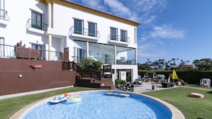 Una casa de dos pisos con piscina, terraza de madera y jardín cercado.