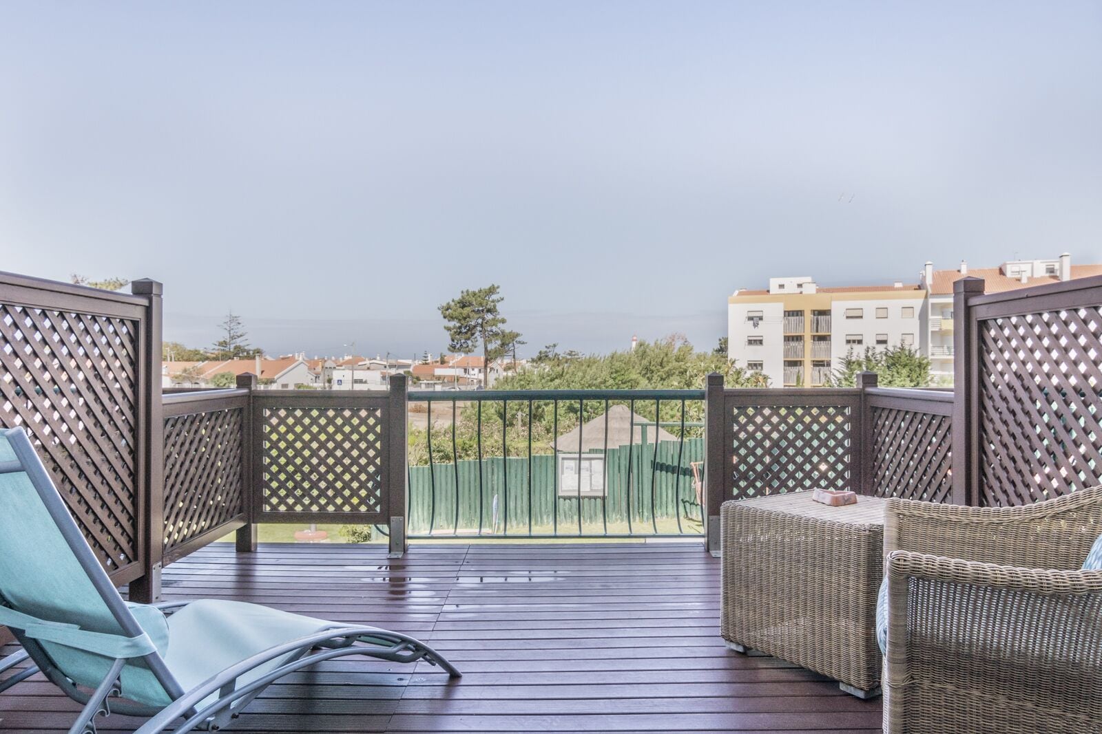 A balcony with a wicker sofa and a chair, wooden flooring, and a view of buildings and trees.