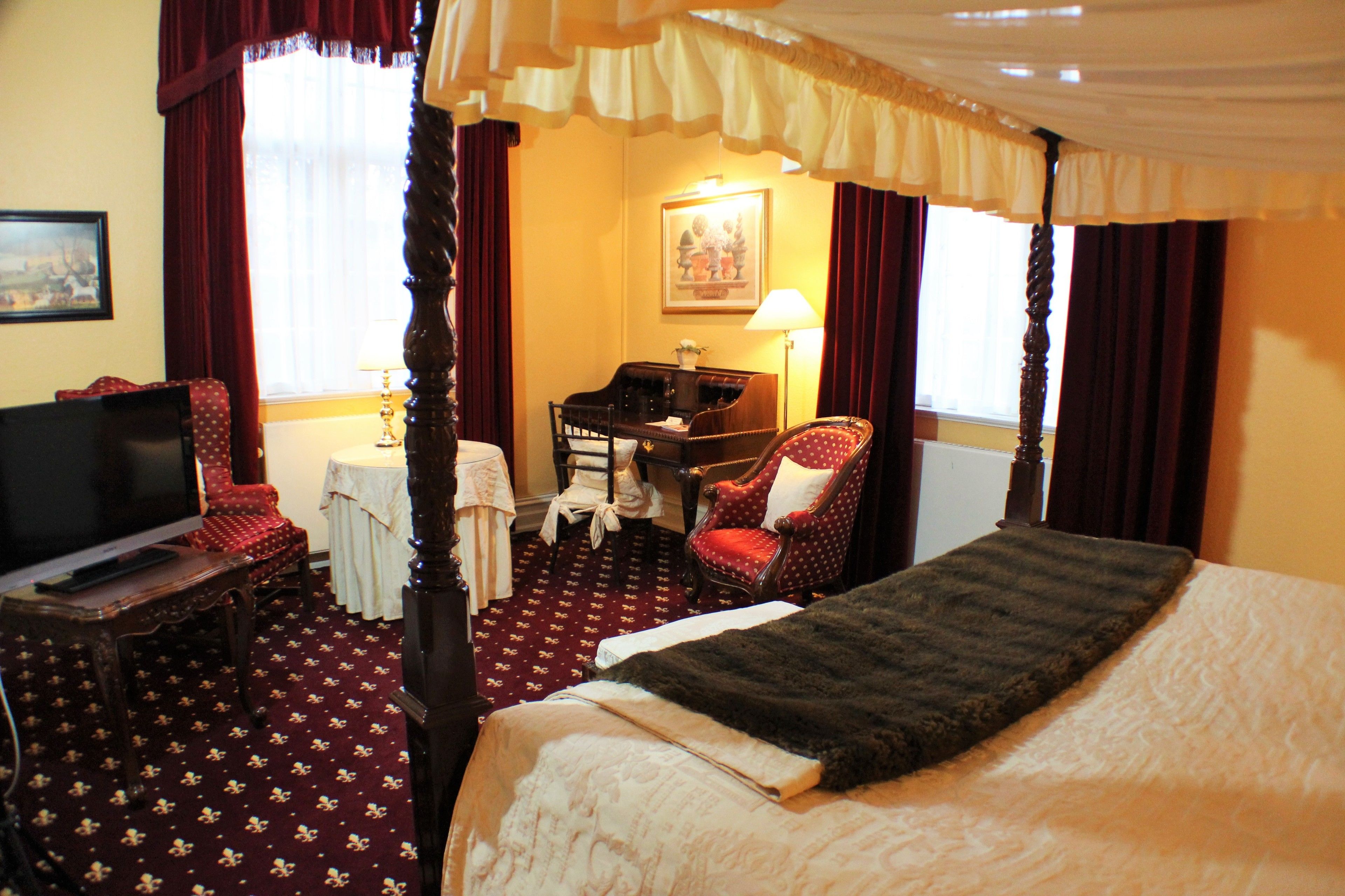 A four-poster bed with a dark throw, a television, a desk with a lamp, and a red polka dot chair.