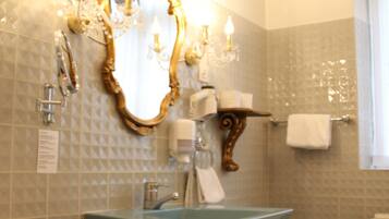 Une salle de bain équipée d’un meuble-lavabo, d’un évier et de toilettes. On y trouve un miroir avec un cadre orné, un porte-serviettes et une table en verre sur laquelle sont posés une bougie et un verre.