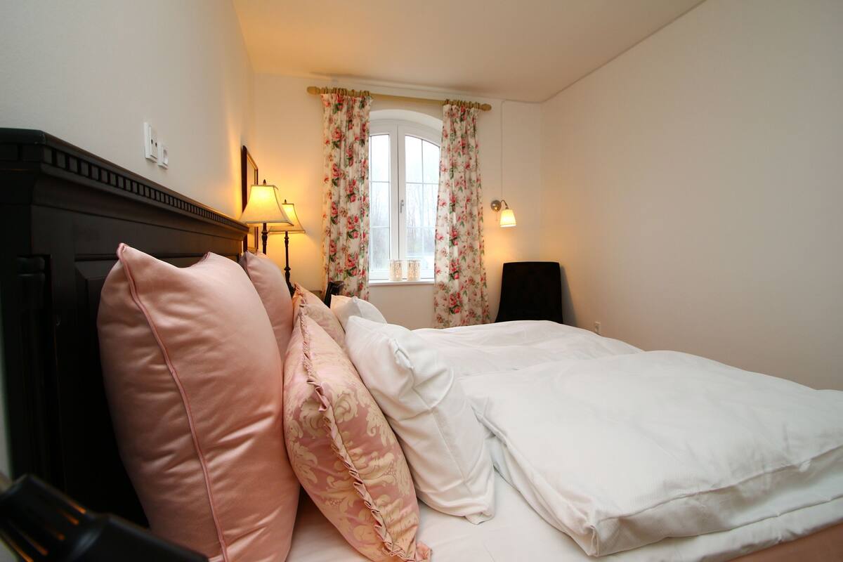 A bedroom with a bed, pillows, a lamp, a window with curtains, and a wall-mounted light fixture.
