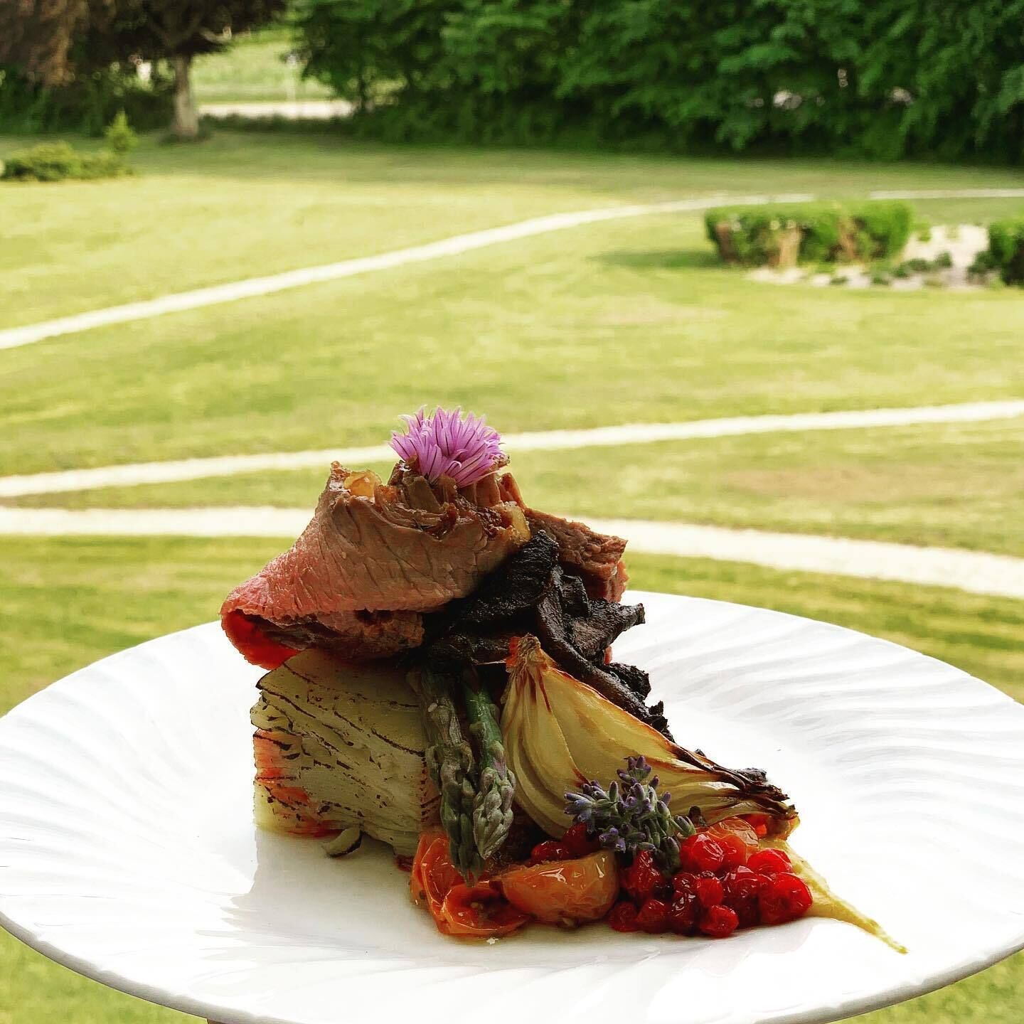 Un plato blanco con diversos alimentos, incluyendo carne, verduras y una flor morada como adorno.