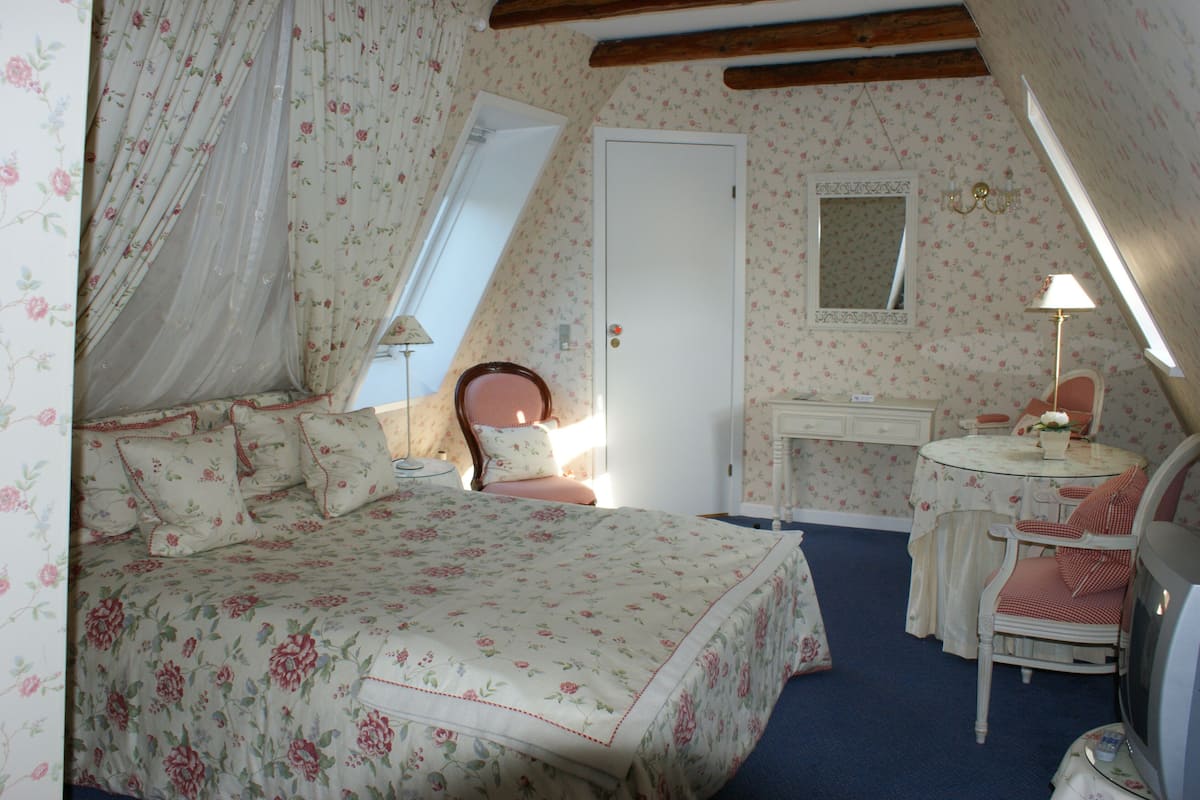 A bedroom with a floral bedspread, a bedside table with a lamp, a chair, and a small table with a vase.