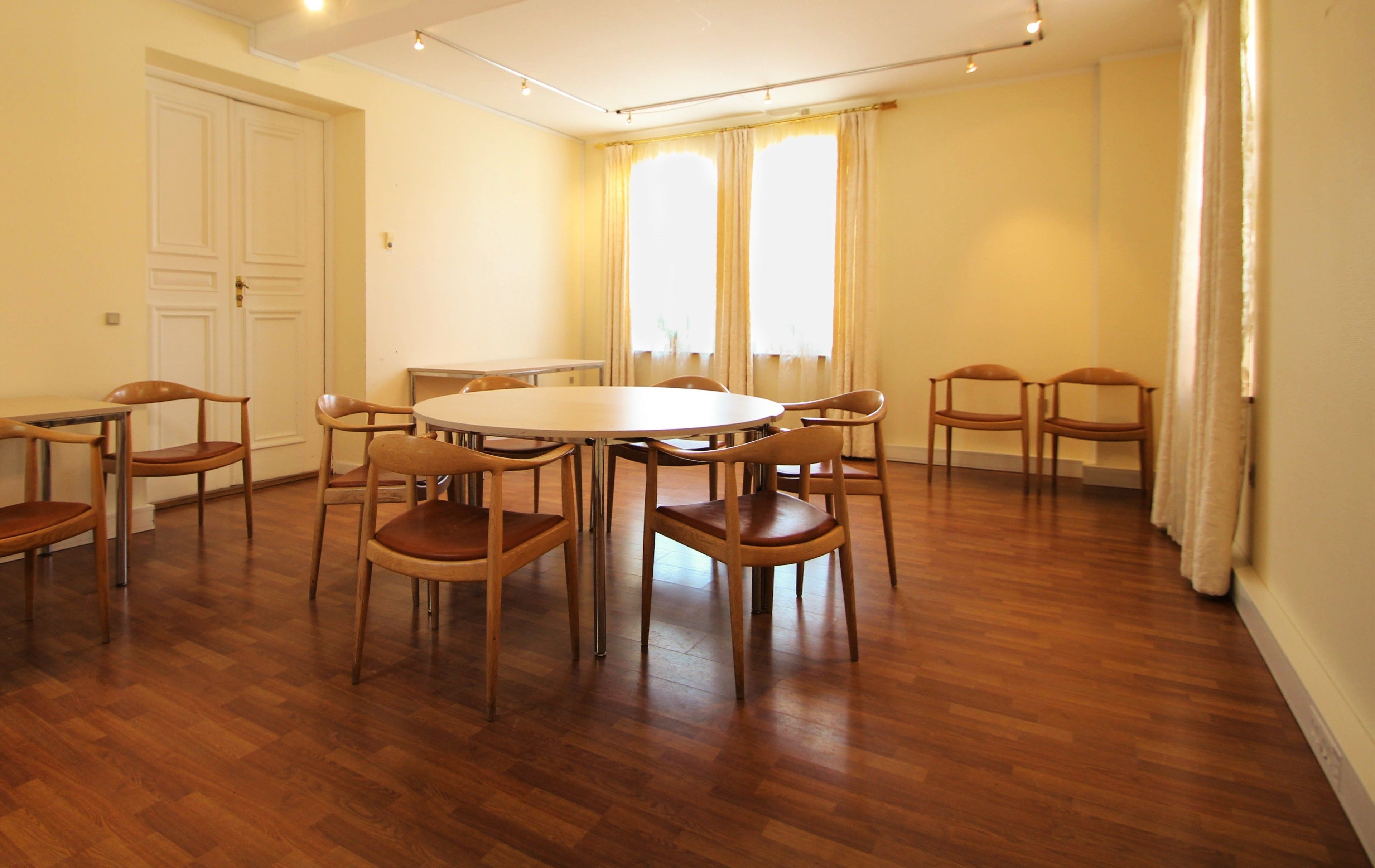 Una habitación amplia con una mesa redonda y ocho sillas de madera, ventanas grandes con cortinas y una puerta.