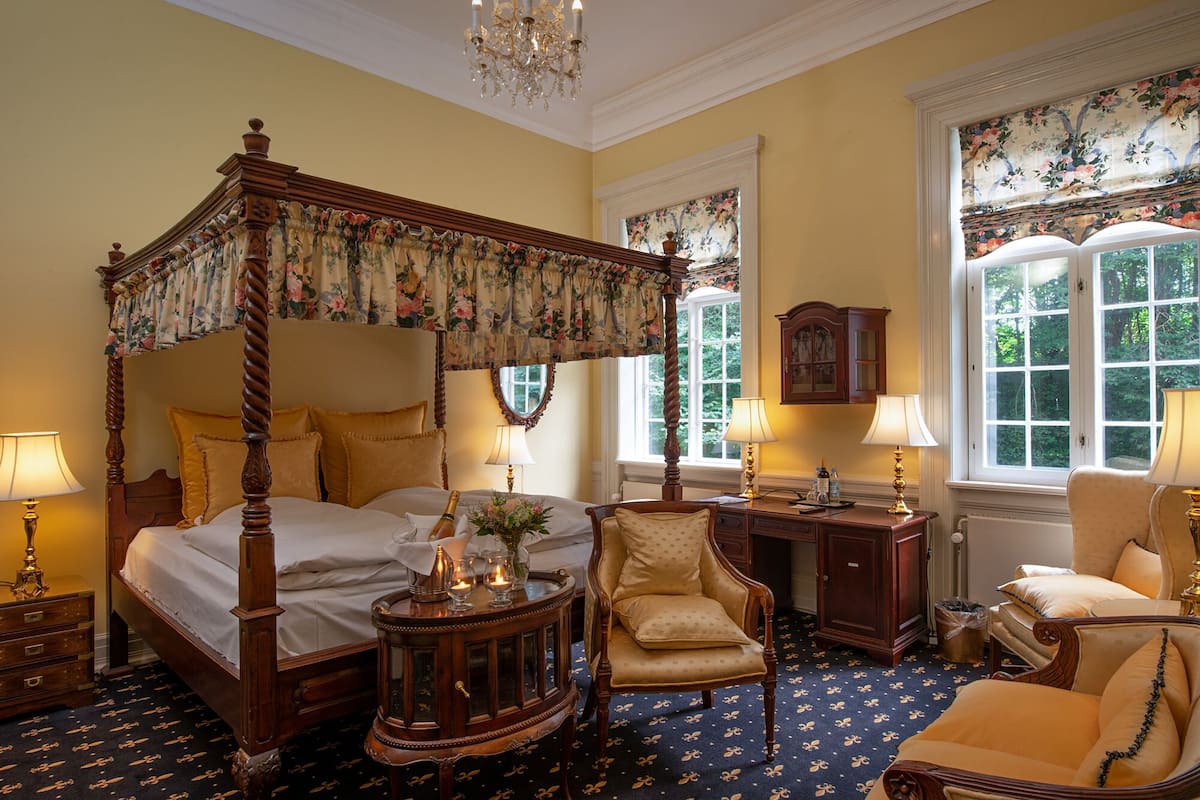 A four-poster bed with floral curtains, a wooden desk, two chairs, and a chandelier.