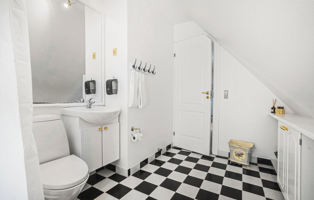 Een badkamer met een wit toilet, wastafel en spiegel. Handdoeken hangen aan haken en er staat een afvalbak. De vloer heeft een zwart-wit geruit patroon.