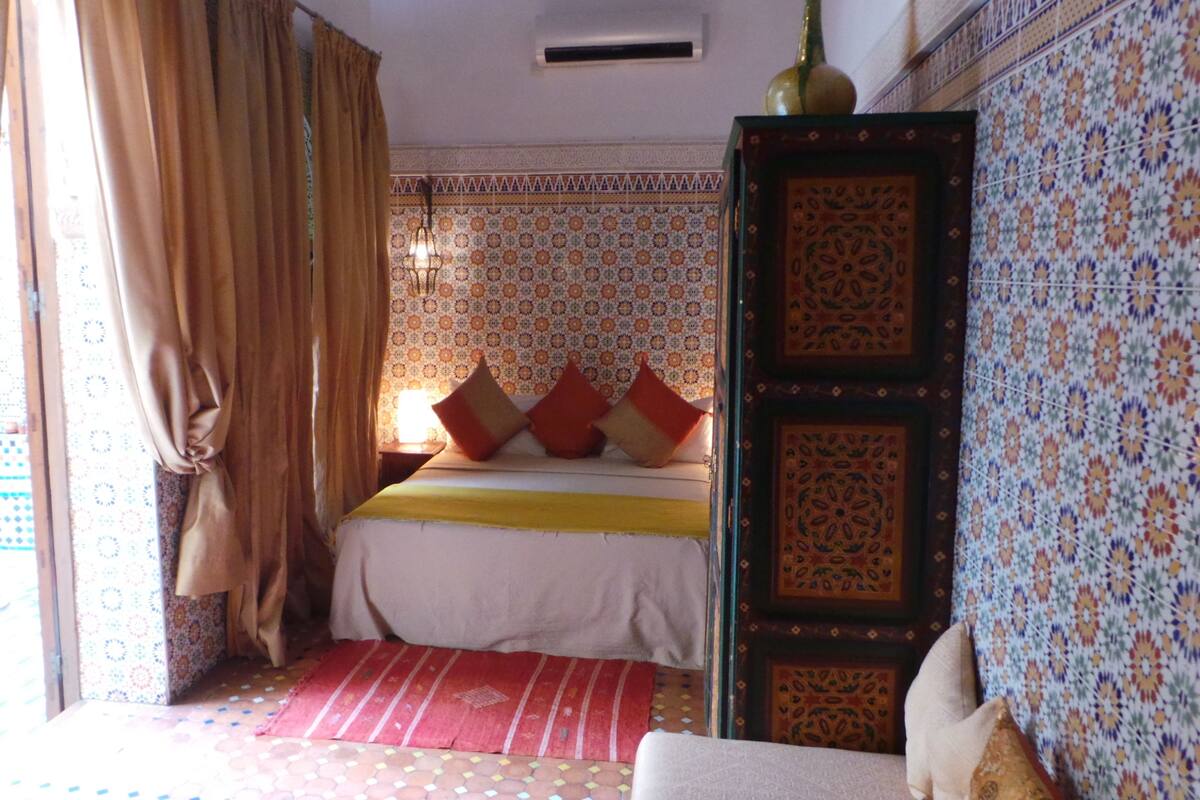 A bedroom with a bed, curtains, patterned walls, and a tiled floor.