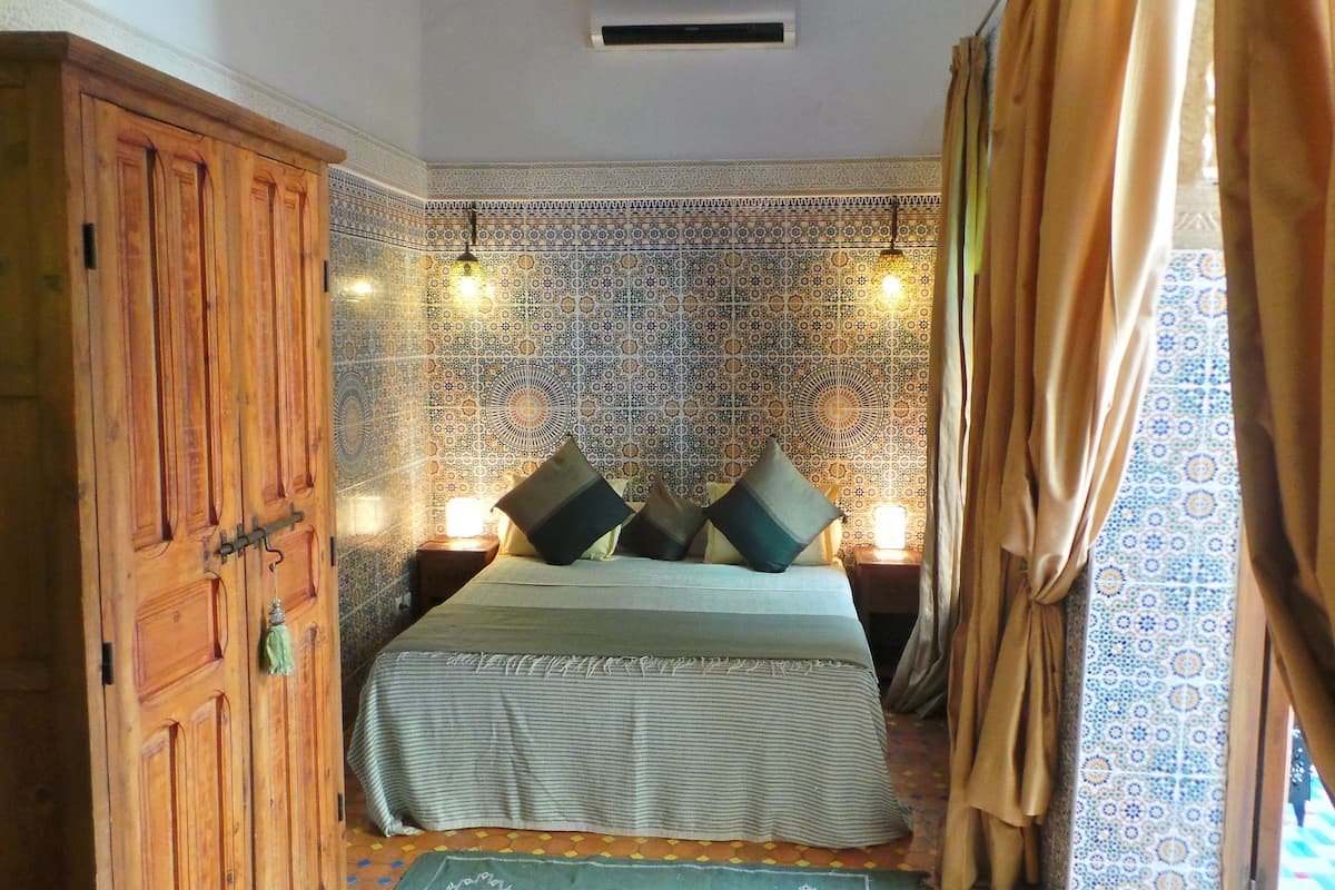A bedroom with a bed, pillows, a wooden wardrobe, patterned walls, and a hanging light fixture.