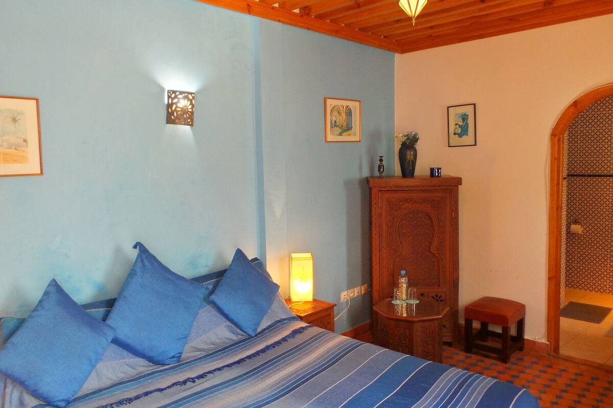 A bedroom with a bed, pillows, a nightstand, a lamp, a wooden cabinet, and a doorway leading to another room.