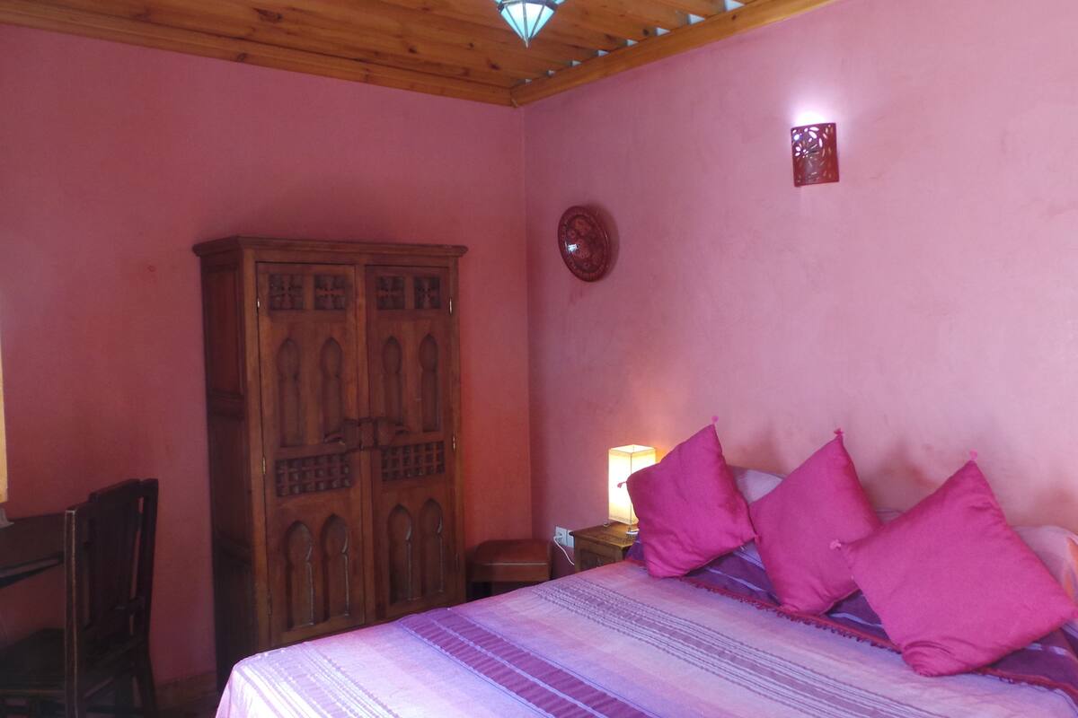 A bedroom with a wooden wardrobe, a bed with pink pillows, and a nightstand with a lamp.
