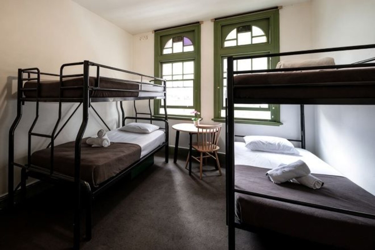 A room with bunk beds, a small round table with a chair, and two windows with green frames.