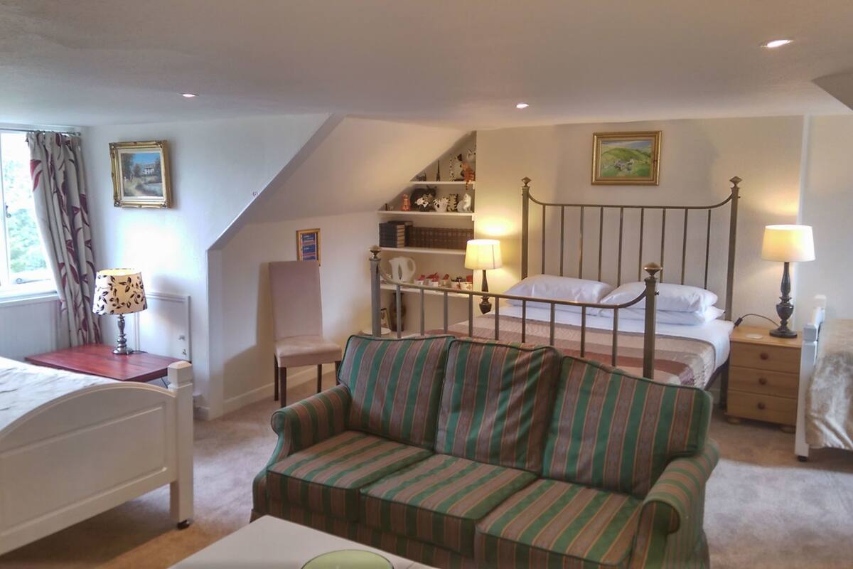 A bedroom with a bed, a striped sofa, a nightstand, a lamp, a window with curtains, a bookshelf, and a painting on the wall.