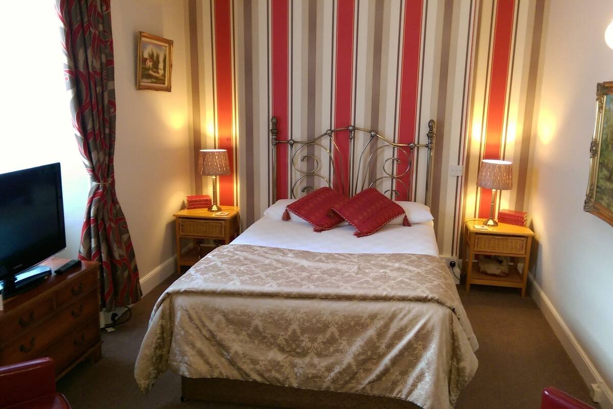 A hotel room with a bed, two bedside tables with lamps, a television, a dresser, and a painting on the wall.
