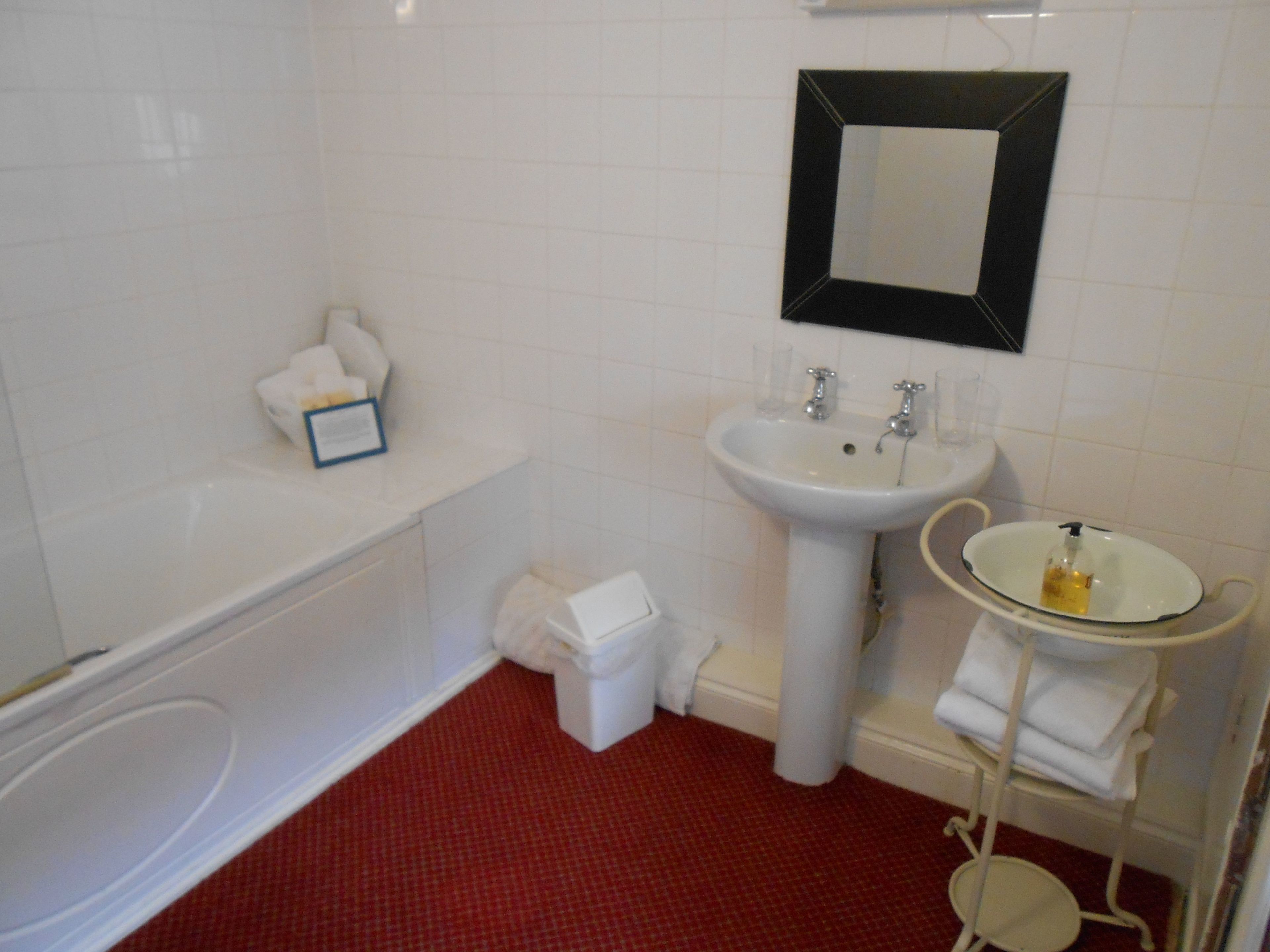 A bathroom with a bathtub, a sink, and a mirror. There is a towel rack with towels and a trash bin.