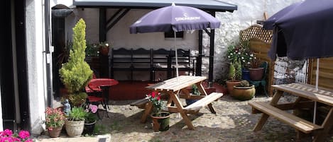 Un patio con un banco de madera, una mesa y un paraguas morado. Hay plantas en macetas y una pequeña estatua.