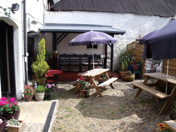 Un patio con un banco de madera, una mesa y un paraguas morado. Hay plantas en macetas y una pequeña estatua.