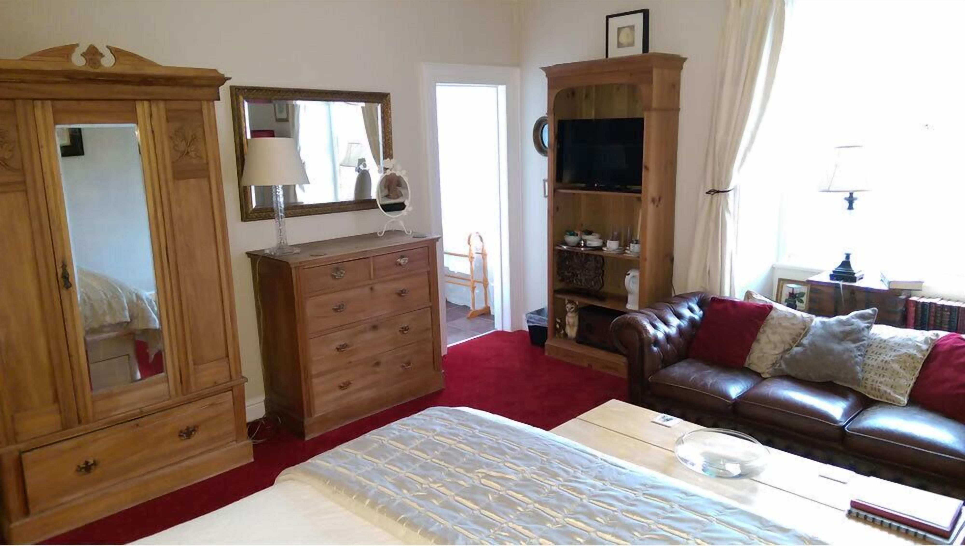 Un dormitorio con un armario de madera, una cama con cubrecama estampado, un sofá de cuero con cojines y un televisor sobre un mueble de madera.