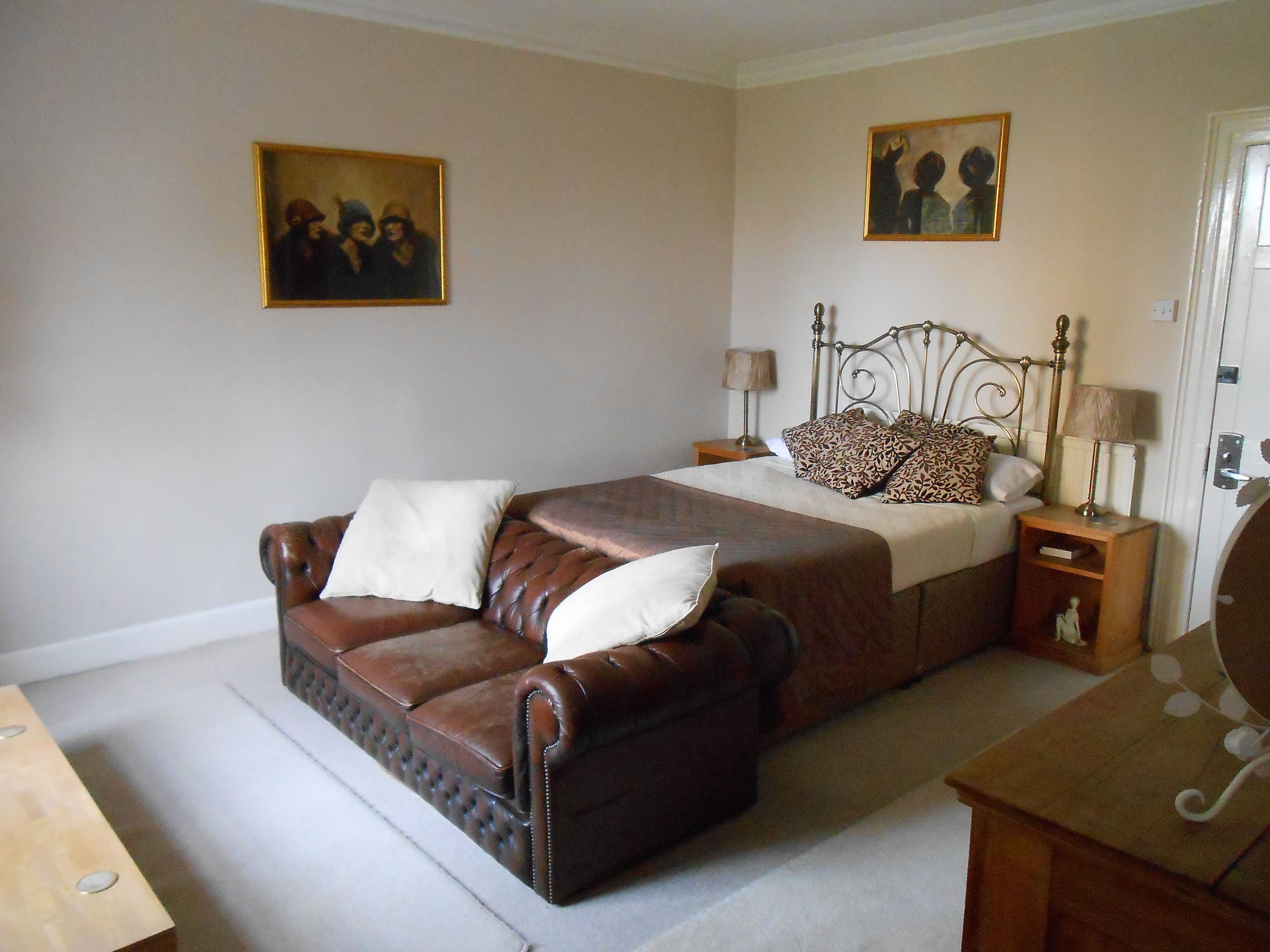 Een slaapkamer met een leren bank, een bed met gekleurde kussens, twee nachtkastjes met lampen, en twee ingelijste foto’s aan de muur.