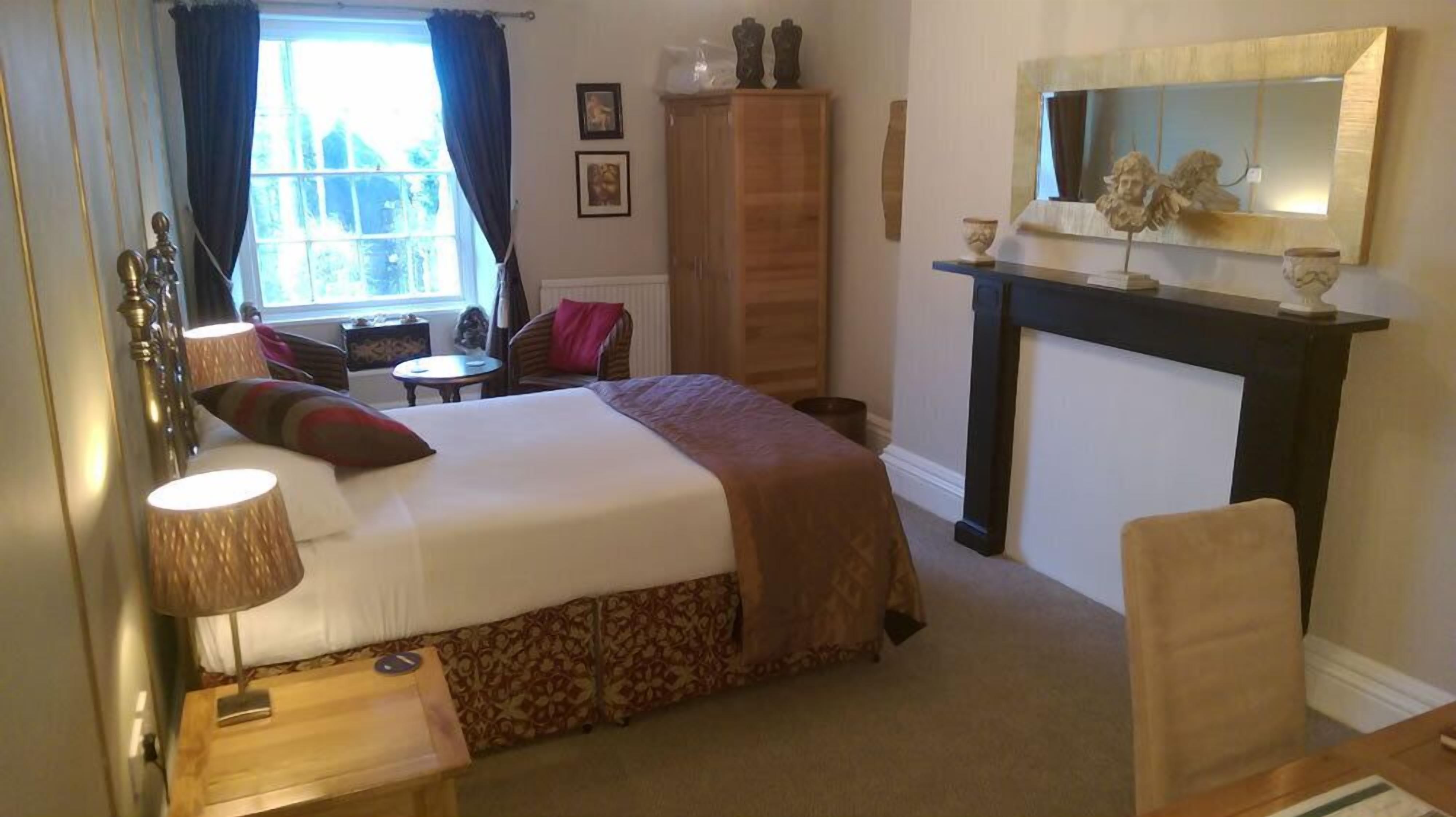 A bedroom with a bed, bedside table, lamp, wardrobe, chair, and a fireplace.
