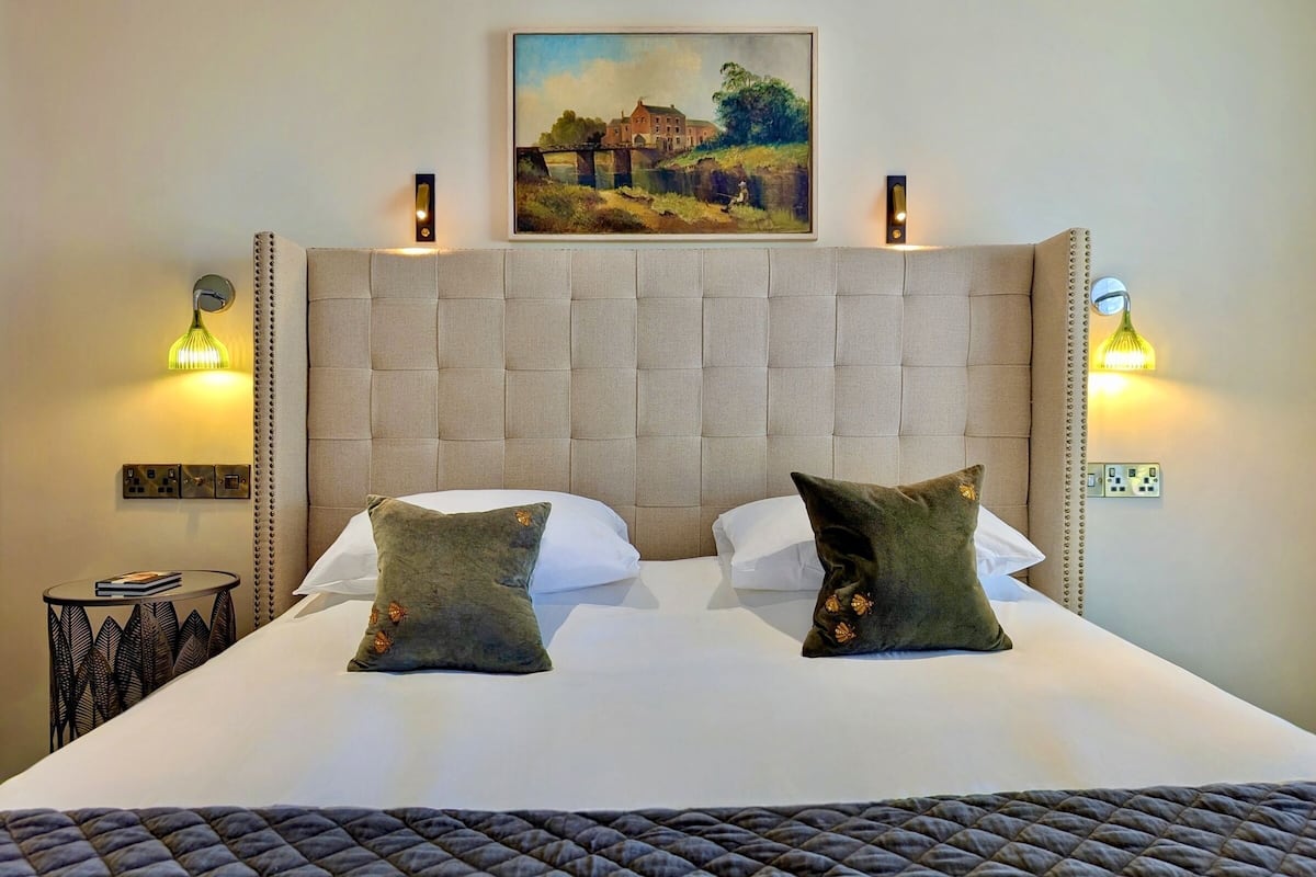 A neatly made bed with two pillows, a headboard with a painting above, and wall sconces on either side.
