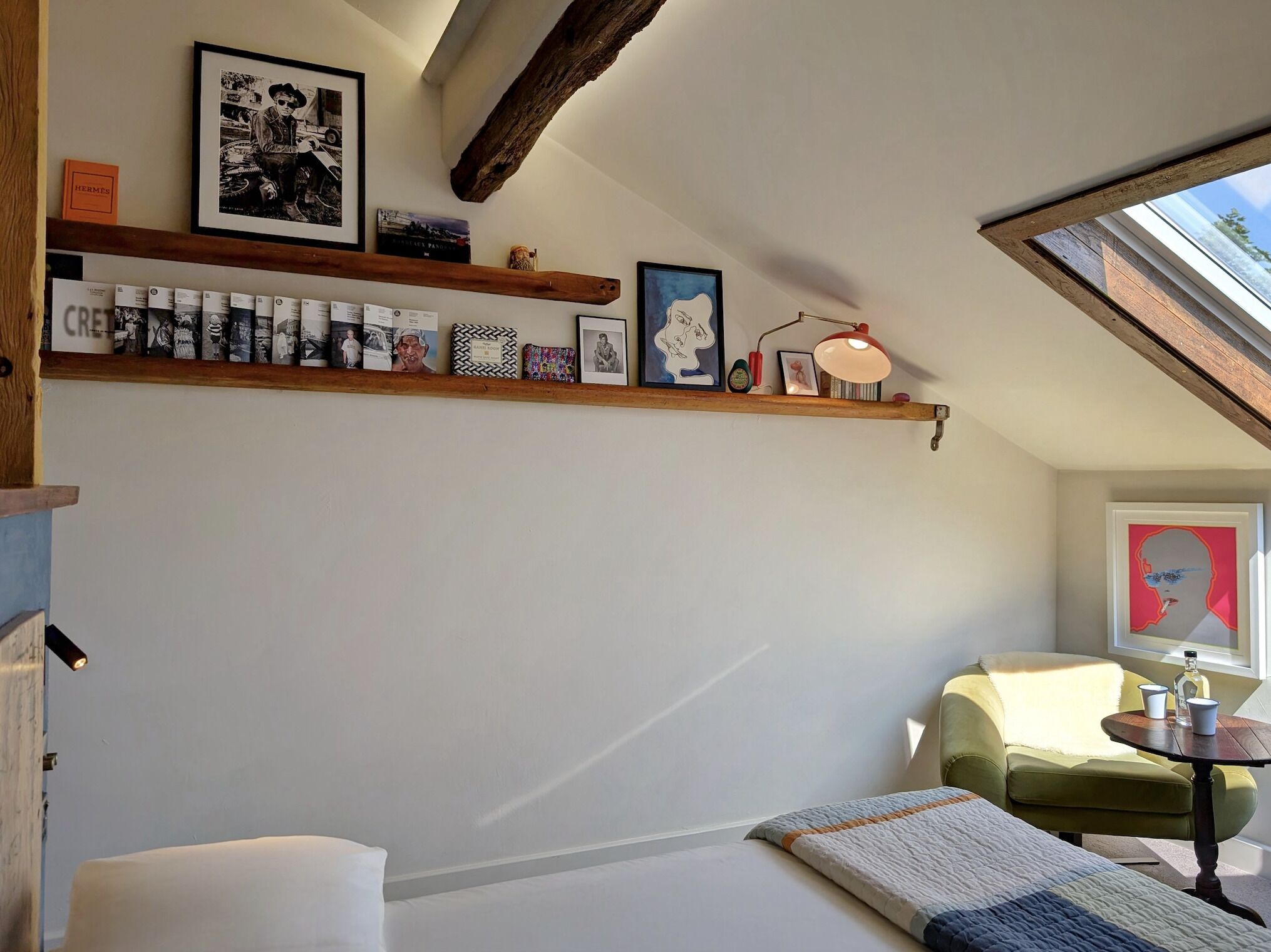 A cozy room with a sloped ceiling, a wooden shelf with various items, a green armchair, and a small round table with a bottle and cups.