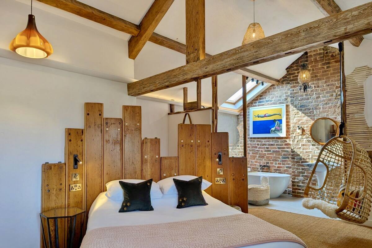 A bedroom with a wooden headboard, a bed with two black pillows, a hanging chair, and a brick wall with a painting.