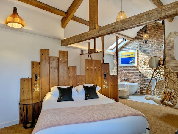 Un dormitorio con cabecera de madera, cama con dos almohadas negras, una silla colgante y una pared de ladrillo con un cuadro.