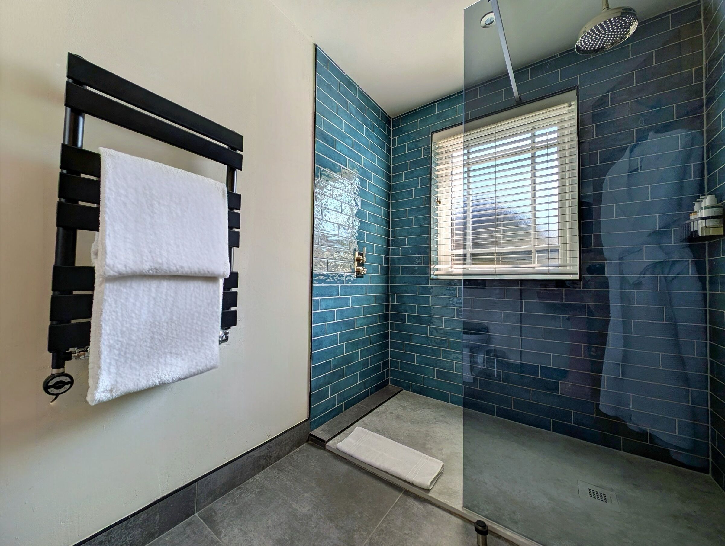 A bathroom with a glass shower enclosure, a towel rack with two towels, and a window with blinds.