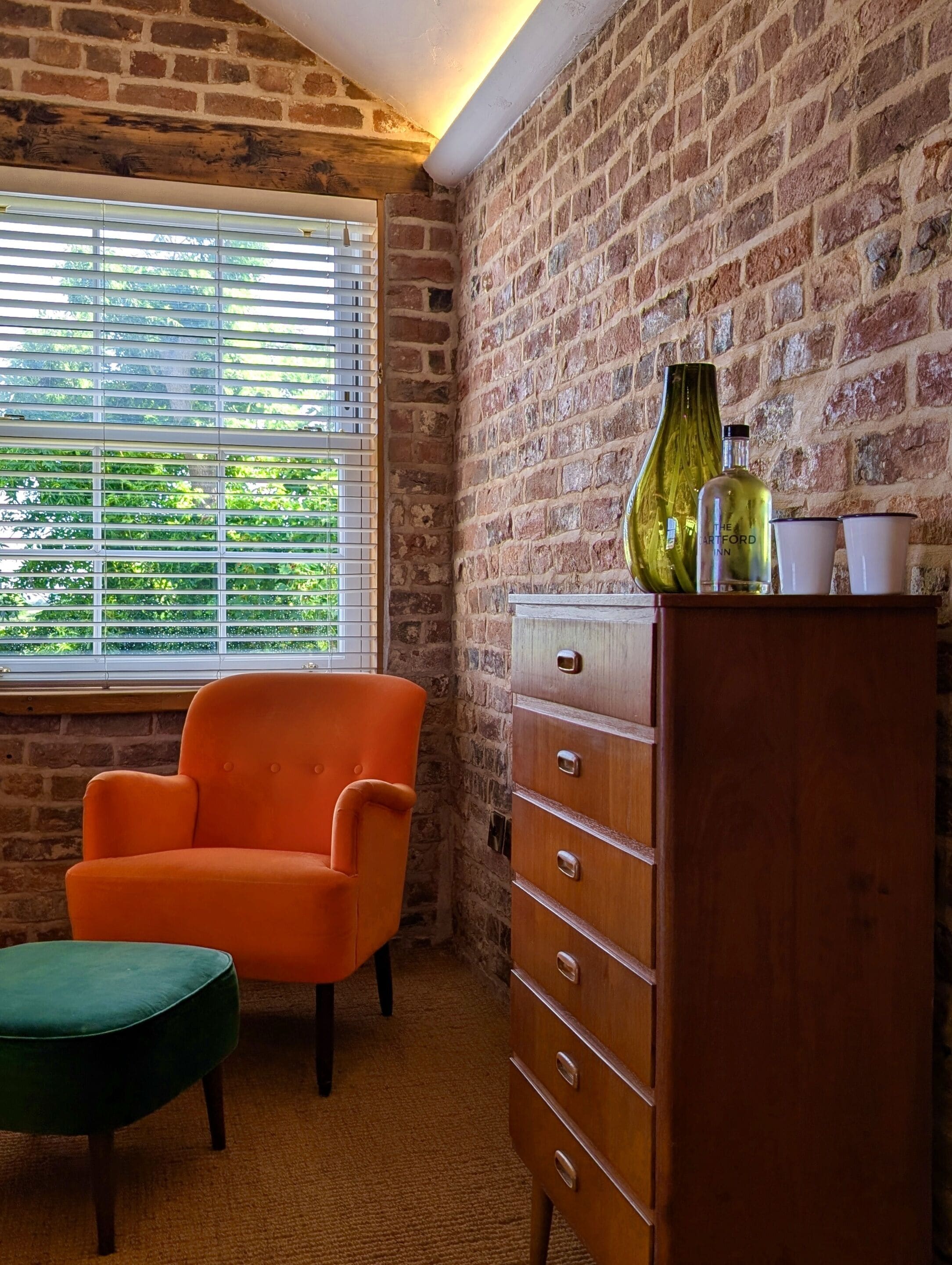 Una habitación con pared de ladrillo, ventana con persianas, una silla naranja, un reposapiés verde y un aparador de madera con una botella y vasos encima.