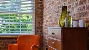 A room with a brick wall, a window with blinds, an orange chair, a green ottoman, and a wooden dresser with a bottle and cups on top.