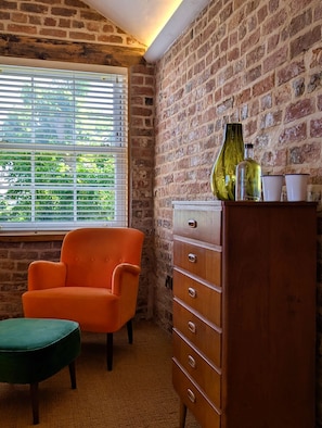 A room with a brick wall, a window with blinds, an orange chair, a green ottoman, and a wooden dresser with a bottle and cups on top. - The Cartford Inn (Preston)